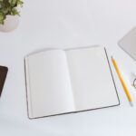 An open empty notebook on a white desk next to an iPhone and a MacBook