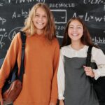 Two smiling students with backpacks standing in front of a chalkboard filled with math formulas.