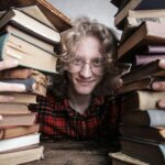 A young man in glasses surrounded by stacks of vintage books indoors, embodying knowledge and learning.