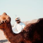 A man in traditional attire posing with camels in a desert landscape.
